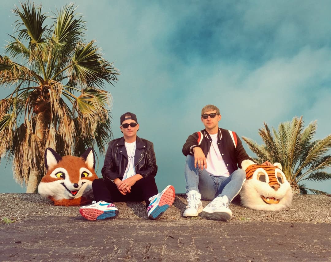 two guys sitting in front of a sky with a palm trees with two animal heads next to them